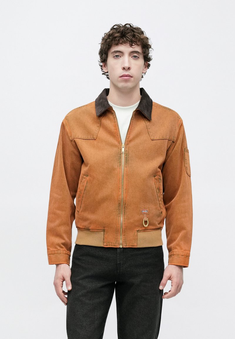 Brown zip-front jacket with a dark collar, two side pockets, ribbed hem, and a metal ring detail. Textured fabric in a relaxed fit.