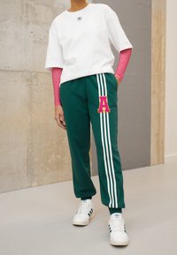 Zelené teplákové kalhoty s bílými pruhy, růžový dekorativní písmeno "A", v kombinaci s bílým tričkem a dlouhým růžovým tričkem pod ním. Bílé tenisky.