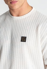Weißer gerippter Pullover mit Strickstruktur und einem kleinen schwarzen Quadrat-Logo mit einem goldenen "G"-Akzent auf der Vorderseite.