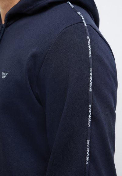 Μπλε σκούρο φούτερ με υφή στο ύφασμα, με ασημένιο λογότυπο στο στήθος και λευκή λωρίδα με λογότυπο κατά μήκος του μανικιού.