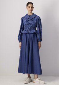 Blusa blu a fantasia con collarino oversize e maniche voluminosi, abbinata a una gonna blu fluente. Il modello indossa scarpe da ginnastica bianche.