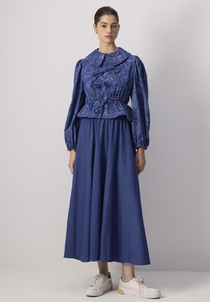 Sininen kuvioitu paita, jossa on ylisuuri kaulus ja puffihihat, yhdistettynä valuvan siniseen hameeseen. Mallilla on päällään valkoiset tennarit.