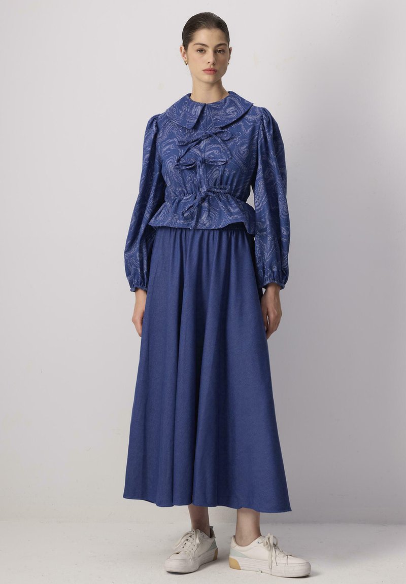 Blusa blu a fantasia con collarino oversize e maniche voluminosi, abbinata a una gonna blu fluente. Il modello indossa scarpe da ginnastica bianche.