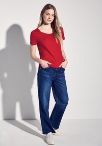 Rød kortærmet T-shirt i afslappet fit, parret med mørkeblå, løstsiddende jeans. Hvide sneakers fuldender outfittet. Minimalistisk indstilling.