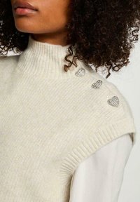 Maglione bianco lavorato a maglia con colletto alto a coste e tre decorazioni a forma di cuore argentate sulla spalla. Texture morbida.