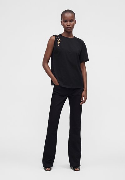 Femme portant un top asymétrique à manches courtes noir, un pantalon noir et des talons ouverts noirs, debout avec une main dans la poche.