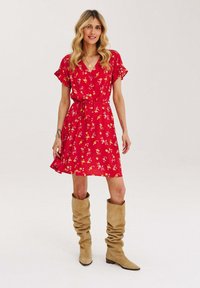 Czerwona sukienka w kwiaty z krótkimi rękawami i przewiązanym w pasie, zestawiona z wysokimi beżowymi botkami w stylu slouch. Materiał wydaje się lekki i ma zwiewny fason.