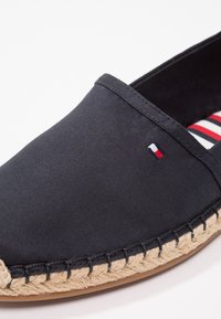Chaussure à enfiler en tissu noir avec une semelle en jute, présentant un logo tricolore et une doublure en textile rayé. Texture lisse et design simple.