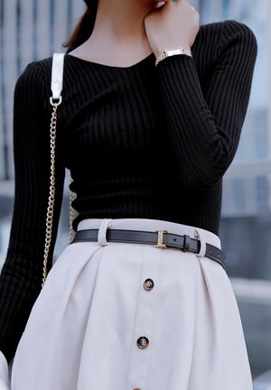 Frau trägt ein schwarzes, geripptes Langarmshirt, einen beigefarbenen High-Waist-Rock mit Knöpfen, schwarzen Gürtel, goldene Uhr und eine weiße Handtasche mit Kettenriemen.