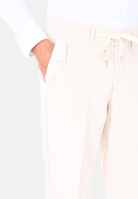 Pantaloni beige chiaro con vita elastica, chiusura con coulisse, tasche laterali e dettaglio della tasca a toppa con bottone. Tessuto morbido.