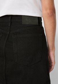 Svarte dongeri med en matt finish, utstyrt med et grønt lærmerke i livet og standard baklommer med sydd detaljer.