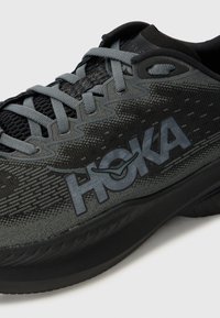 Zwarte sportieve schoen met een bovenwerk van mesh, grijze accenten en het HOKA-logo. Beschikt over een gevoerde zool en een gestructureerd oppervlak voor grip.