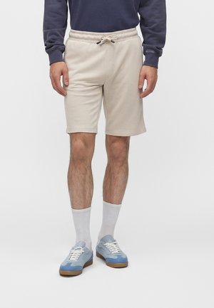 Mann trägt beige Shorts mit Kordelzug, weiße Sportsocken, hellblaue Sneaker und ein dunkelblaues Langarmshirt und steht vor einem weißen Hintergrund.