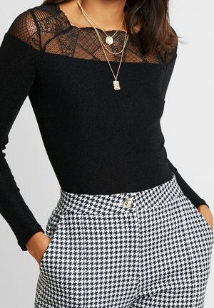 Mujer con blusa negra de manga larga con escote de encaje y collares dorados en capas, combinada con pantalones de pata de gallo en blanco y negro de cintura alta.