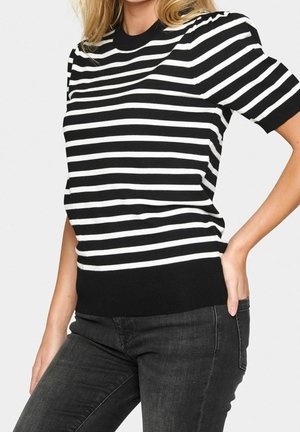 Kobieta nosząca czarno-biały, poziomo prążkowany top z krótkim rękawem w połączeniu z ciemnoszarymi dżinsami, pozująca z jedną ręką na biodrze.