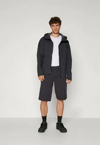 Crna vodootporna jakna s kapuljačom, s patentnim zatvaračima; uparen s tamnim šortsevima, crnim čarapama i cipelama, na neutralnoj pozadini.