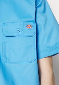 Lichtblauw kortarmshirt gemaakt van stevig materiaal, met een borstzak met een achthoekige klep en een logopatch.