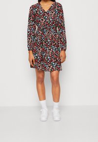 Das Blumenkleid hat einen schwarzen Hintergrund mit roten, blauen und grünen Blumen. Es hat lange Ärmel, einen V-Ausschnitt, eine taillierte Taille und eine Länge über dem Knie.