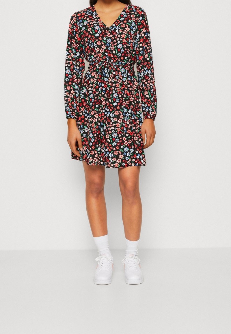 Das Blumenkleid hat einen schwarzen Hintergrund mit roten, blauen und grünen Blumen. Es hat lange Ärmel, einen V-Ausschnitt, eine taillierte Taille und eine Länge über dem Knie.