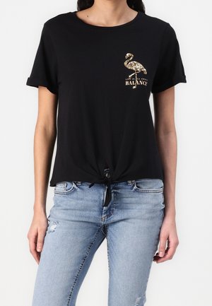 Mujer con top negro de manga corta con un flamenco de lentejuelas doradas y el texto "La vida se trata de equilibrio", combinado con jeans azul claro desgastados.