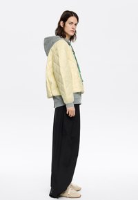 Gequiltede gele jas over een grijze hoodie, met groene hardwareaccenten, zwarte loszittende broeken en beige schoeisel.