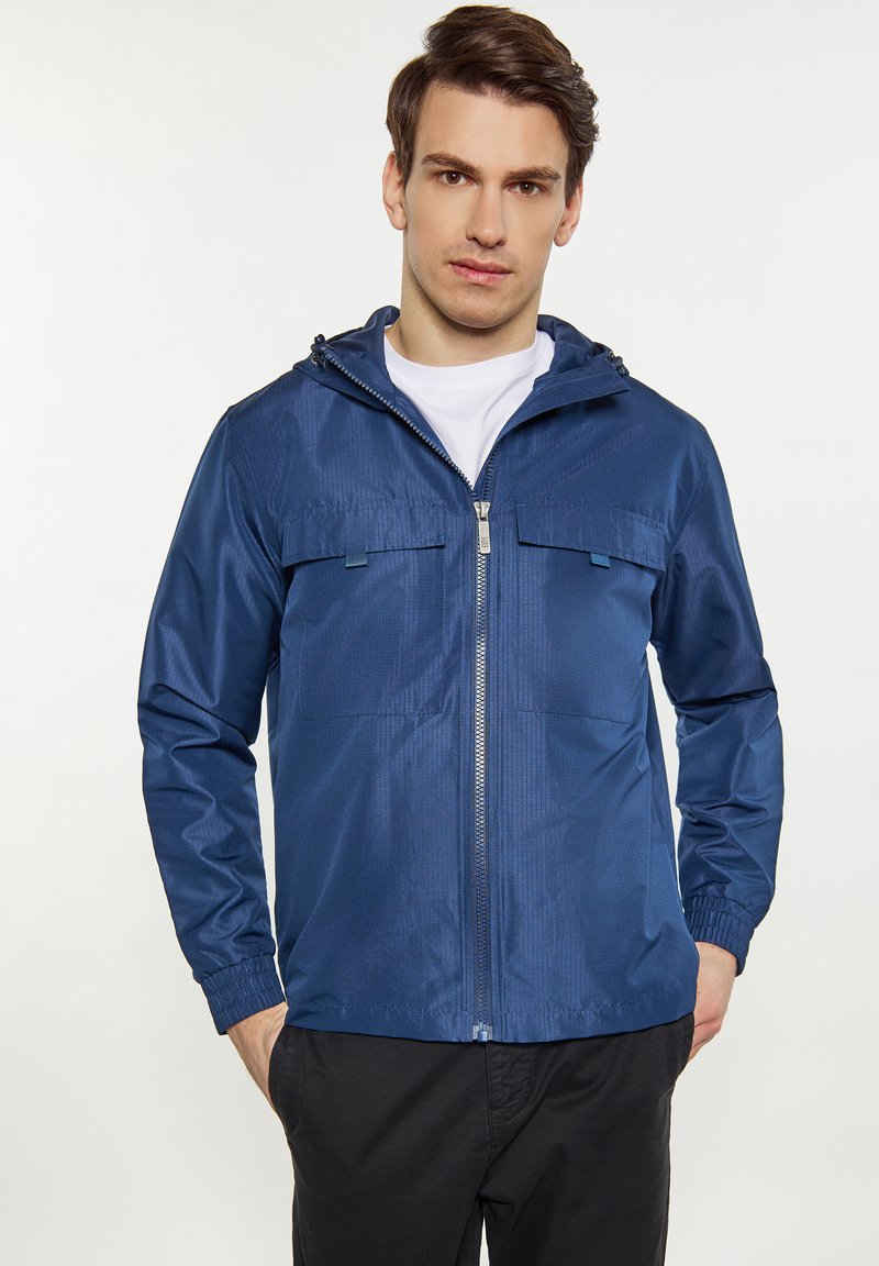 Tamsiai mėlynas lengvas striukė su užtrauktuku, kapišonu ir dviem krūtinės kišenėmis; pagaminta iš lygios, blizgios medžiagos su elastingomis rankovėmis.