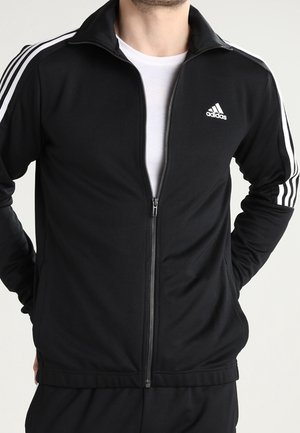 Homme portant une veste de survêtement Adidas noire avec des bandes blanches sur les manches et le logo Adidas sur la poitrine, zippée à mi-hauteur par-dessus une chemise blanche.