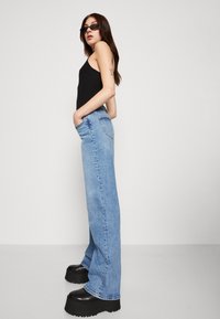 Črn rebrast top, svetlo modri široki jeans, chunky črne platform čevlje. Model stoji bočno ob nevtralni ozadju.