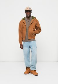 Brun hættejakke med grøn foring, grå polo, lyseblå jeans og brune støvler. Robust design med synlige syninger og lommer.