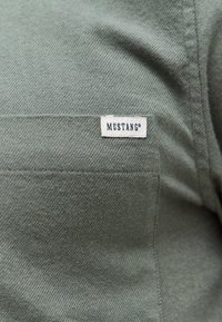 Zielony materiał z ukośnym prążkowanym wzorem. Na tkaninie wszyta jest biała metka z napisem "MUSTANG®".