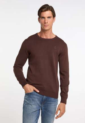 Mann trägt einen braunen Pullover mit langen Ärmeln und blaue Jeans, eine Hand in der Tasche, steht vor einem schlichten weißen Hintergrund.