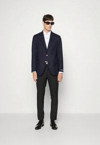 Jūrinės mėlynos spalvos švarkas su auksiniais mygtukais, balta marškinėlių apykakle, juodos kelnės ir juodi batai. Modelis dėvi juodus saulės akinius.