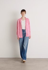 Pink, textured jacket over a white top, paired with light blue, loose-fitting jeans. Model stands in front of a plain background.