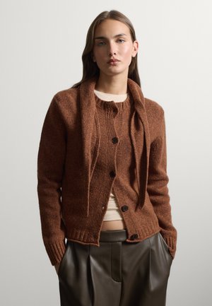 Junge Frau trägt eine braune Strickjacke mit passendem Schal und dunkle hoch taillierte Lederhose, steht mit den Händen in den Taschen vor einer weißen Wand.
