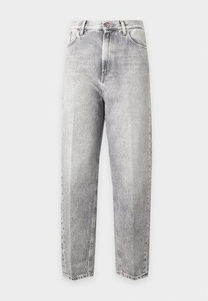 Jeune personne portant un haut blanc sans manches drapé, un jean gris clair et des ballerines noires pointues, debout avec une main dans la poche.