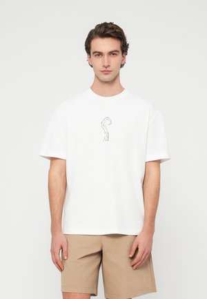 Bărbat în picioare, purtând un tricou alb cu un mic design negru în forma unui tentacul de caracatiță pe piept și pantaloni scurți bej, pe un fundal simplu.