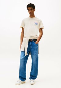 Creme t-shirt med et farverigt grafisk design, løse blå denimjeans, sort bælte, beige strikket sweater om livet og hvide sneakers.