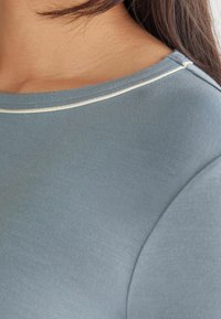 Ljusblå tajt topp gjord av mjukt material, med rundad halsringning och creamfärgad accentstickning längs kragen. Slät textur.