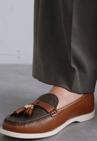 Bruine leren loafers met tekstuur van zwart stof, een gouden kettingaccent en een witte rubberen zool, gedragen met grijze stoffen broek.