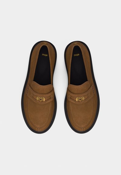 Mocassins à enfiler en daim marron avec semelles noires et emblème décoratif doré sur les brides, vus de dessus sur fond blanc.