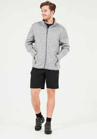Grå zip-up jacka med en texturerad yta, två sidofickor, bärs över en marinblå skjorta, tillsammans med svarta shorts och svarta skor med gröna detaljer.