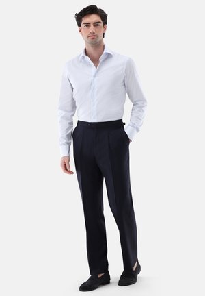 Mann steht, trägt ein hellblau gestreiftes Hemd, hochtaillierte dunkle Hose und schwarze Loafers vor einfachem Hintergrund.