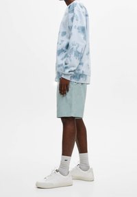 Sweat-shirt tie-dye bleu clair avec poignets côtelés, associé à un short vert clair. Le mannequin porte des baskets blanches et des chaussettes blanches montantes à la cheville.