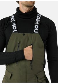 Groene waterdichte overalls met verstelbare zwarte banden, voorzien van de tekst "MOU" en met ritszakken. Draag ze over een zwarte geribbelde top.