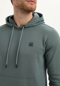 Man in een grijs-groene hoodie met een voorzak en trekkoorden, die een deel van zijn baard en nek toont tegen een witte achtergrond.
