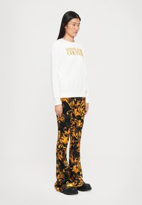 Felpa bianca con logo "VERSACE JEANS COUTURE" in oro, abbinata a pantaloni neri con un vivace motivo a spirale giallo e arancione.