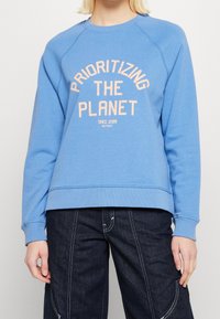 Ljusblå sweatshirt i mjukt tyg, med orange texten "PRIORITIZING THE PLANET" i bågformad design. Ribbad krage och ärmslut.