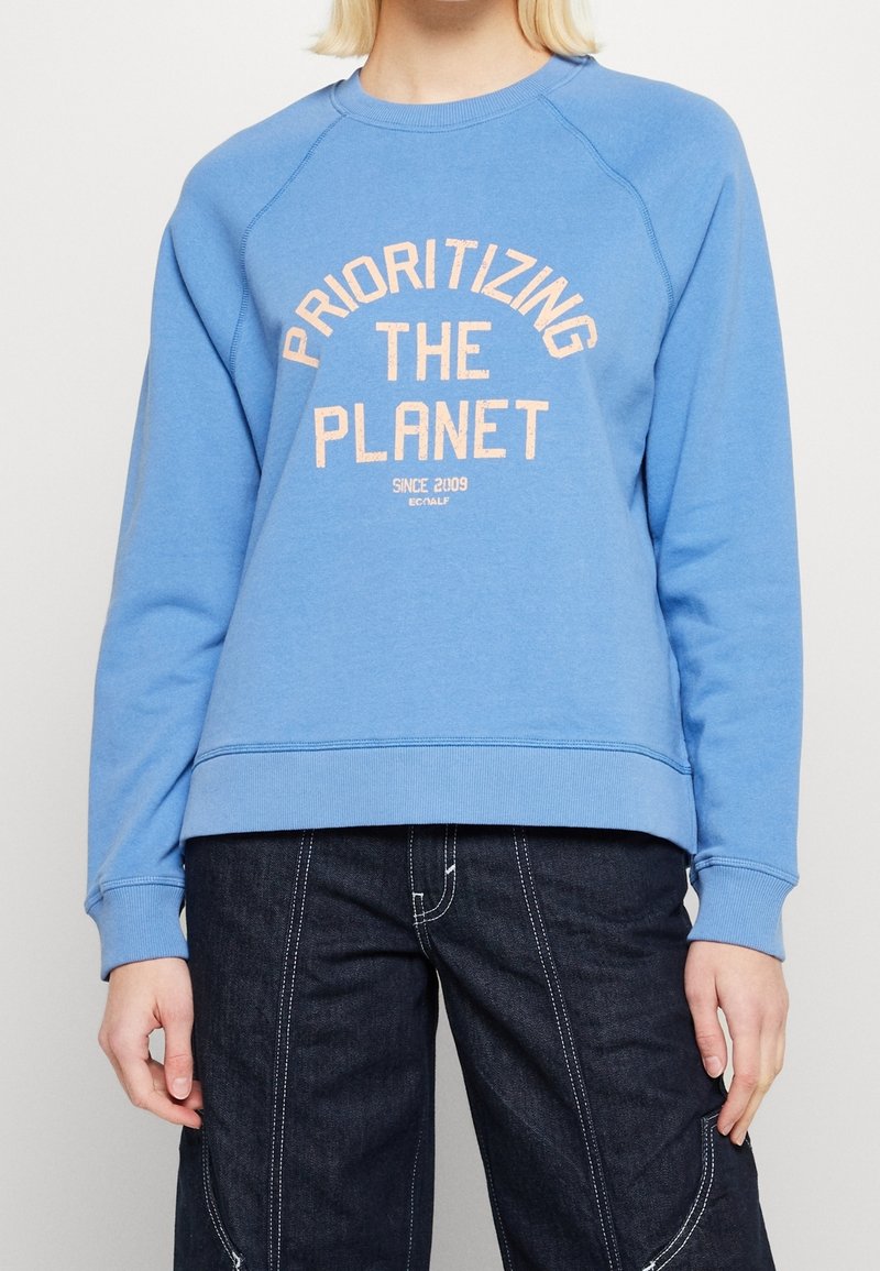 Ljusblå sweatshirt i mjukt tyg, med orange texten "PRIORITIZING THE PLANET" i bågformad design. Ribbad krage och ärmslut.