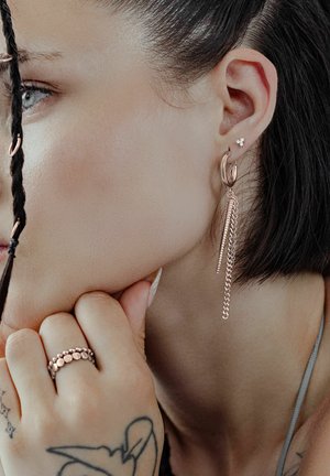 Woman's profile with braided hair, multiple earrings including a long chain earring, hand with a ring, and a visible tattoo on the wrist.