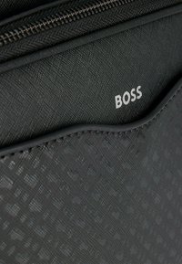 Portafoglio nero con texture, chiusura a zip e motivo embossato. Presenta il logo "BOSS" in argento su una superficie liscia.